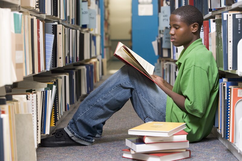 Male university student sitting on library floor surrounded by books